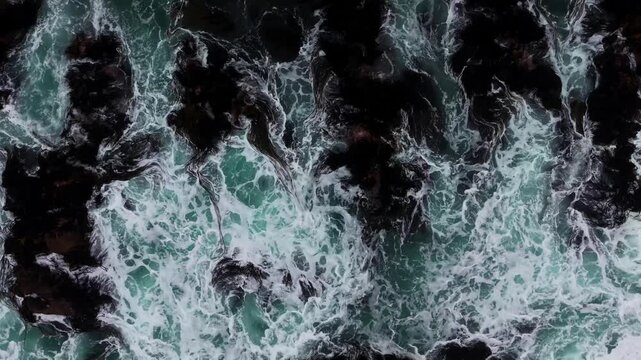 Drone Horizontal Stationary Top Down View of Ocean Waves Crashing on Dark Rocky Shore with Seaweed, Chile