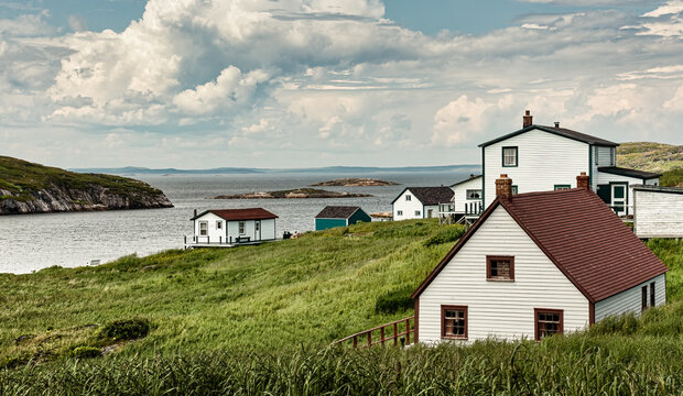 Weathered wooden houses and fishing sheds line a grassy hillside above a rocky cove at Battle Harbour, a historic fishing settlement on the Labrador coast. Scattered clouds fill the sky above calm wat