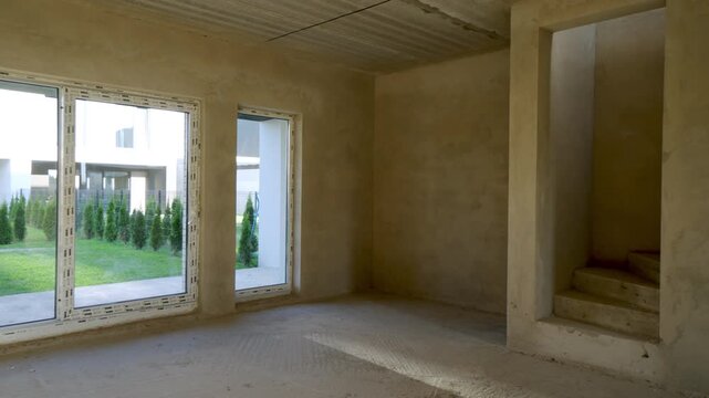 Unfinished house construction showing interior space with concrete walls, panoramic windows overlooking a green backyard and stairs leading to the next floor