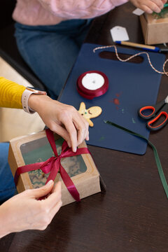 Diverse female friends tying red satin ribbon on kraft window box at home table, packaging cookies