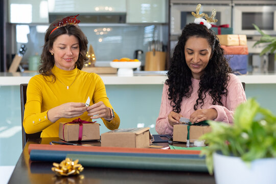 Two female friends wearing holiday headbands wrapping gifts at home kitchen with brown boxes