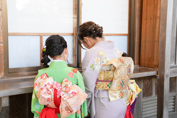 Obraz premium Two women in traditional Japanese kimonos stand beside a wooden window. One wears a vibrant green kimono with floral patterns. The other dons a soft lavender robe with embroidered designs