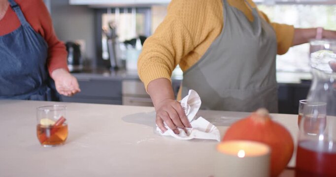 Diverse female friends with aprons wiping kitchen island after baking, clearing flour with pitcher