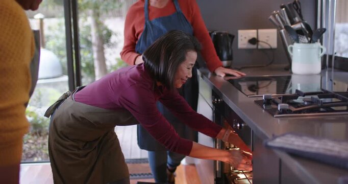 Diverse females with senior in aprons responding to timer opening oven and removing tray at counter