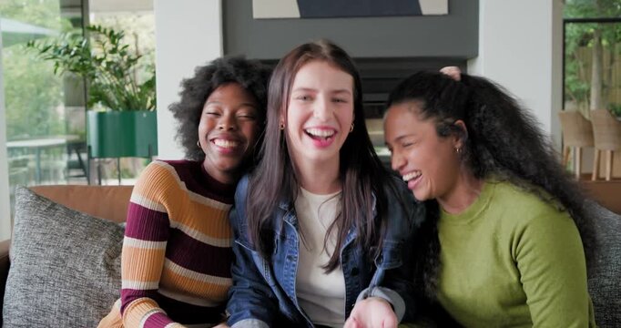 Diverse female friends laughing and leaning on sofa in living room reacting to right friend comment
