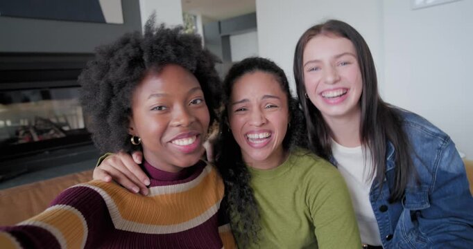 Three female friends extending phone, posing, taking selfie and laughing on couch at home