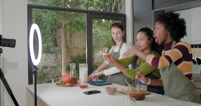 Diverse female friends reacting as ring light signaling filming at kitchen island, drizzling pastry