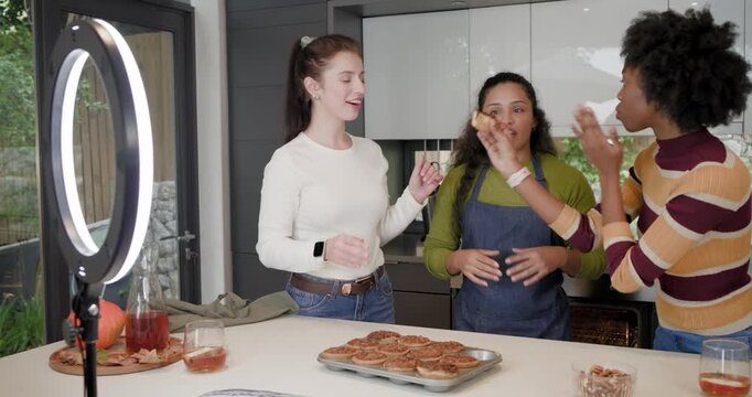 Diverse female friends celebrating fresh bake, offering pastries to ringlight rig at kitchen island