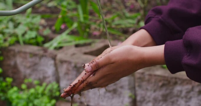 Woman wearing maroon sweatshirt holding hands under thin water stream, rubbing soil from fingers