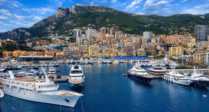 ScreenshotElevated panoramic view of Port Hercule in Monte Carlo, Monaco, showcasing luxury yachts, the vibrant marina, and the city's iconic skyline set against the dramatic Maritime Alps. One of the