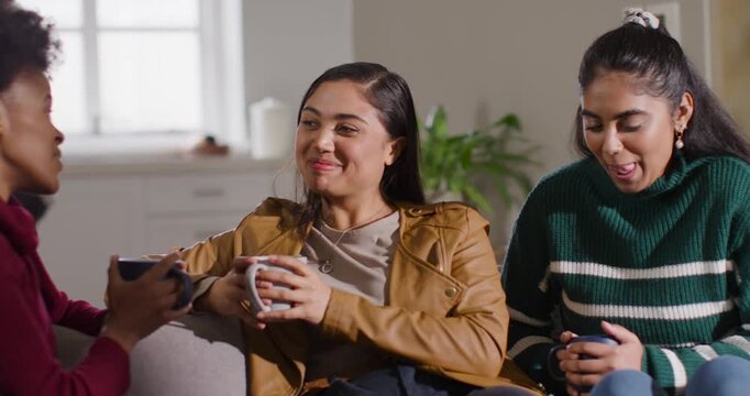 Diverse females sitting on couch holding mugs, central woman reacting to left friend in tan jacket