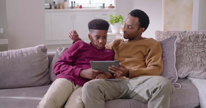 African American father and teenage son leaning on gray sofa, son swiping tablet, father guiding