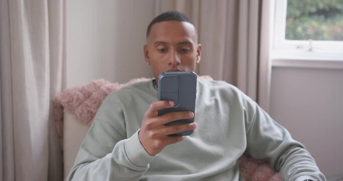 After ping African American man checking phone, scrolling, adjusting in armchair with pink throw