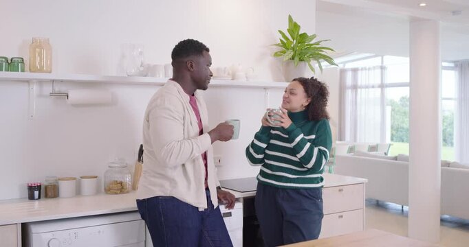 African American couple lifting mugs, sipping, chatting and smiling in kitchen with phone and fruit