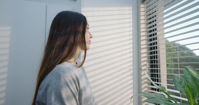 Asian woman pausing while looking through venetian blinds at home, acknowledging camera, smiling