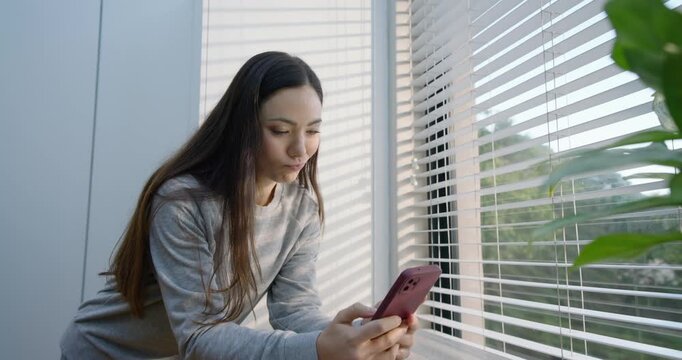 Woman holding pink phone leaning on counter by blinds after text, glancing up, smiling into lens