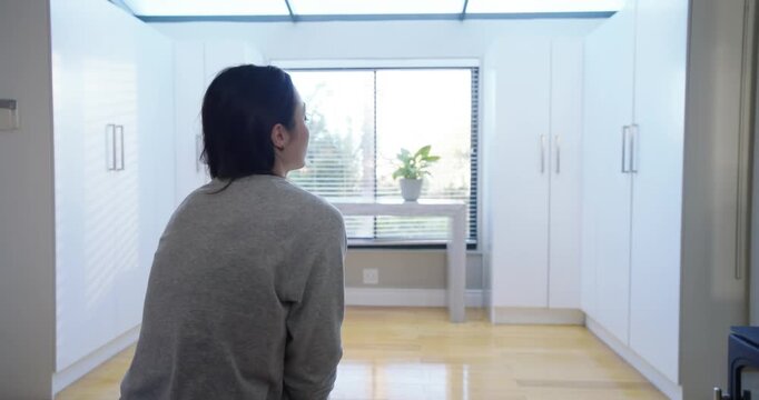 Asian woman wearing gray pajamas, pausing on stool in bright room, straightening toward skylight
