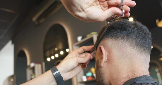 African American barber using comb and scissors cutting client hair at shop, blending fade near ear