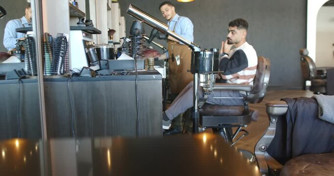 Male barber preparing tools and using hood dryer while male client sitting for styling, copy space