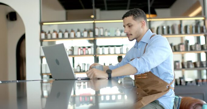 Adult barista brown apron opening laptop, typing on trackpad in cafe sipping cup, checking orders