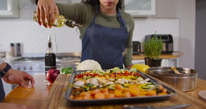 African American woman in apron tilting bottle, drizzling oil over veggies tray at island to roast