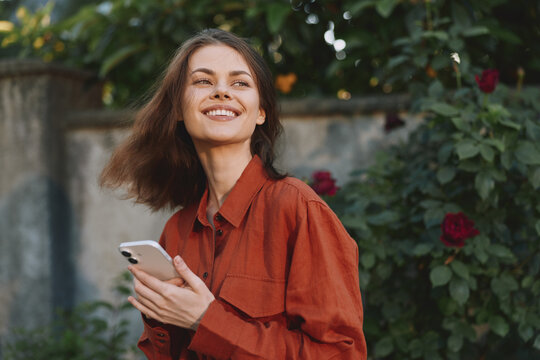 Cheerful woman outdoors, smiling with phone in hand, rust shirt, sunlit garden setting, relaxed casual vibe, natural colors, lush greenery background, in frame today
