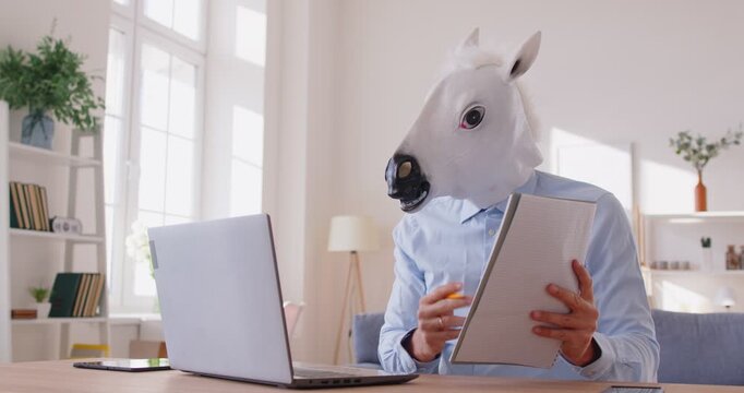 Funny businessman wearing horse head mask is actively participating in online business meeting. In office man uses laptop and gives presentation with clipboard, demonstrating humor and professionalism