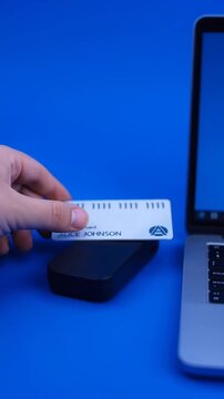Hand holding credit card over contactless payment terminal next to laptop on blue background.