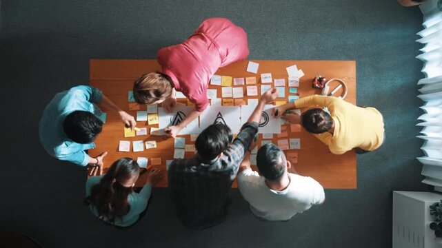 Top down aerial view of skilled designer hold logo while discuss with team. Aerial business group working together sharing idea about logo graphic design by using sticky notes and tablet. Symposium.