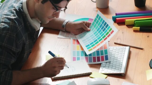 Professional designer writing and designing logo graphic at meeting. Closeup of business man working on colored palettes and writing idea on sticky notes on table with equipment and paper. Symposium.
