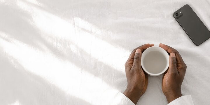 Top view of human hands holding warm beverage cup on morning bed
