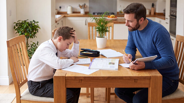 Father sits at a wooden kitchen table helping his young son with school homework while the boy looks frustrated and rests his head on his hand.