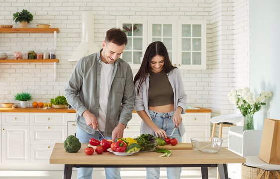 Happy young family couple, cooking salad together, helping, healthy vegans at home modern kitchen, vegetarian diet, vegetables, plate of fresh eco food at table, health benefits of well-balanced food