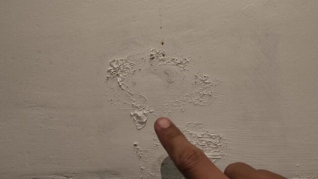 A close up view of a person checking peeling and crumbling white paint texture on a damp concrete wall caused by water leakage.
