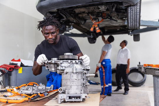 Professional EV technician reassembling an electric drive unit in a modern electric vehicle service center while colleagues inspect a lifted electric vehicle in the background.