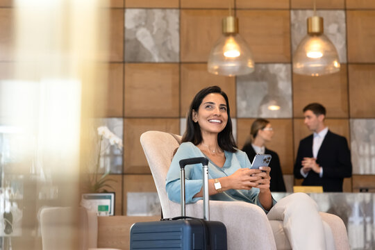 Smiling Latina woman guest using smartphone sitting in hotel lobby with suitcase nearby, waits for taxi or check-in in hotel reception. Travel, hospitality, leisure concept for tourism, accommodation