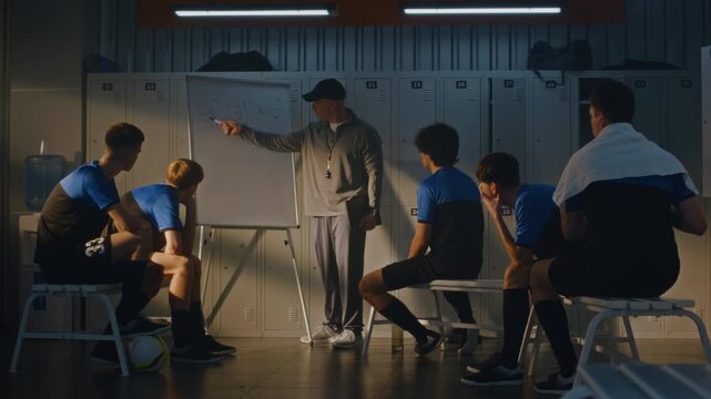 Middle Aged Soccer Coach in Black Cap Stands by Whiteboard, Drawing Game Tactics, Explaining Strategy to Teenage Players. Youth Football Team Sits on Benches in Locker Room, Focused on Presentation.