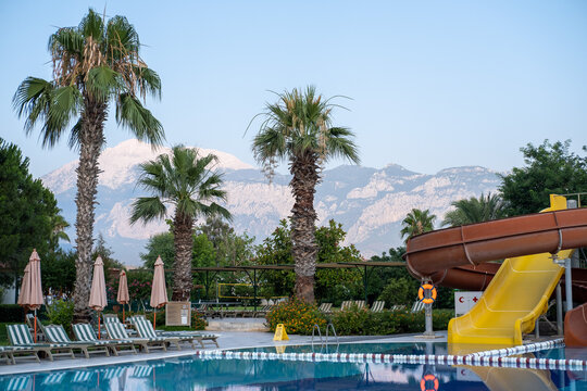 Outdoor swimming pool with colorful water slide, sun loungers and umbrellas at a modern resort hotel in Turkey. Summer vacation destination with mountains in the background and relaxing atmosphere