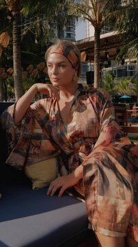 Relaxed woman in patterned beach kimono enjoying sunshine at tropical resort lounge  