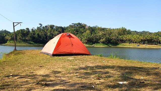 Bright orange camping tent beside a tranquil lake surrounded by lush tropical forest under a clear blue sky.