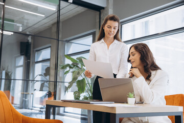 Professional women collaborating, discussing reports and using a laptop in a modern corporate...