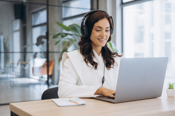 Fototapeta na wymiar Young businesswoman wearing headphones and smiling, typing on laptop while working in a modern office