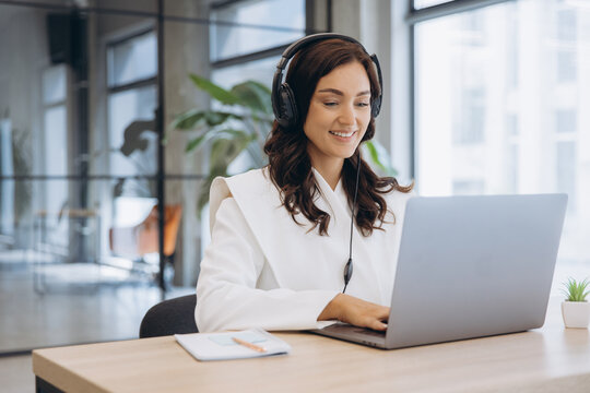 Young businesswoman wearing headphones and smiling, typing on laptop while working in a modern office
