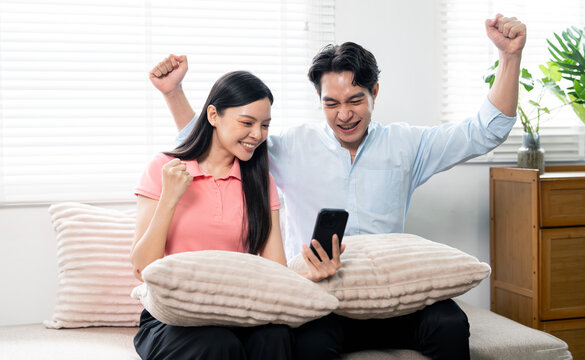 Happy Asian couple celebrating success while looking at smartphone on sofa.