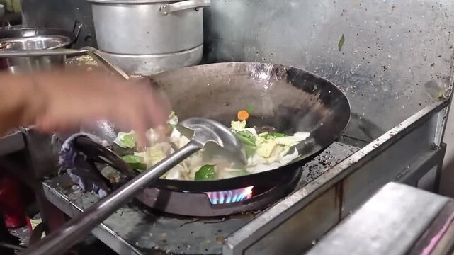 Chef hands stirring fresh mixed vegetables in a large iron wok over a burning gas stove flame.