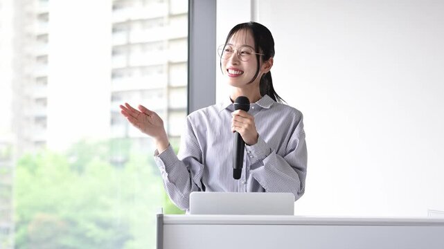 会議室でマイクを持ってプレゼンテーションを行う30代の日本人ビジネスパーソンの女性