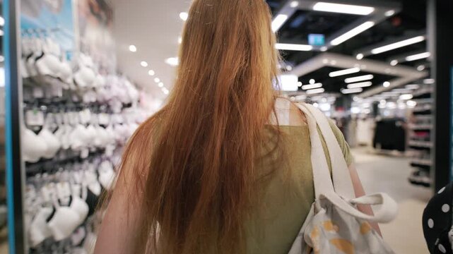 Two women shopping in a mall, one with long red hair and glasses, walking through a clothing store with various bras displayed on racks