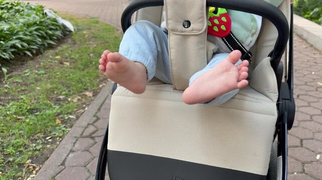 the bare feet of a baby kicking playfully while sitting in a modern beige stroller. A relaxed and happy outdoor family walk on a pleasant day.