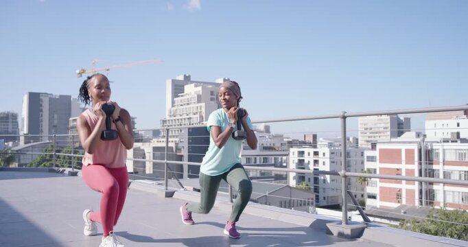 Two African women friends holding dumbbells stepping forward and lunging on rooftop for fitness