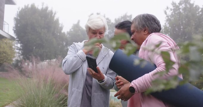 Senior with two women coming to park pink-clad woman holding rolled mat tapping phone planning yoga